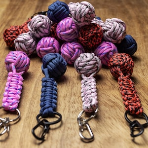 Può includere: Quattro portachiavi colorati con palline annodate e chiusure in metallo. Le palline sono rosa, blu, bianche e rosse con accenti neri e blu. I portachiavi sono su una superficie di legno.