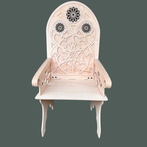 Könnte beinhalten: Ein Holzstuhl mit einer geschnitzten Rückenlehne und filigranen Verzierungen. Der Stuhl hat eine helle Holzoptik und eine einfache, rechteckige Sitzfläche.