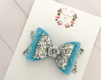May include: A light blue and silver glitter hair bow with a small white flower accent. The bow is on a white card with the text "Daisy Florence Bows" in pink.