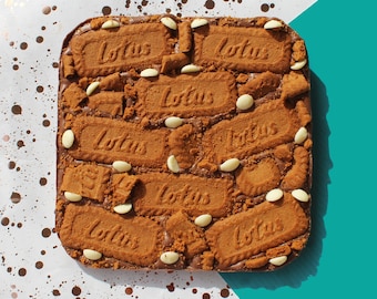 Op de afbeelding: Een chocolade brownie bedekt met Lotus Biscoff-koekjes en witte chocolade chips. De brownie is vierkant en ligt op een wit oppervlak met bruine stippen.