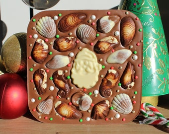 May include: A square milk chocolate bar decorated with seashell-shaped chocolates, a white chocolate Santa Claus, and sprinkles. A green Christmas tree and baubles are in the background.