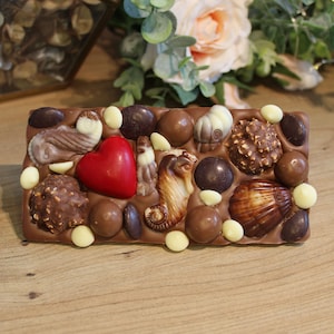 May include: A milk chocolate bar with a variety of toppings. The decorations include a red heart, seashell-shaped chocolates, and round chocolates. The bar is presented on a wooden surface. The bar is approximately 15 cm long.