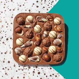May include: A square chocolate slab with an assortment of chocolates. The dark brown base is patterned with lines. The chocolates include white chocolate spheres, shell-shaped chocolates, and chocolate-covered nuts and caramels.