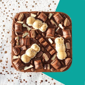 May include: A close-up of a chocolate bar with various toppings, including chocolate pieces, white chocolate, and wafer cookies. The bar is on a white surface with brown spots.