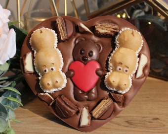 Könnte beinhalten: Eine herzförmige Schokoladenkonfekt mit einem Teddybären, der ein rotes Herz hält, zwei nilpferdförmigen Süßigkeiten und Schokoladen- und Waffelstücken. Die Schokolade hat eine satte braune Farbe, und die Süßigkeiten sind hellbraun und weiß.