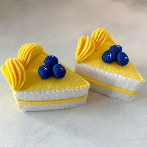 May include: Two felt lemon cakes with white frosting and blue felt blueberries on top. The cakes are yellow and have a swirl of yellow felt on top.