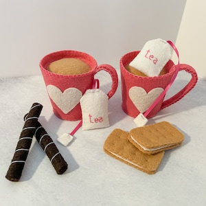 May include: Two pink felt teacups with white felt hearts and tea bags that say "tea" in pink thread. The teacups are on a white surface with two chocolate sticks and two brown and white cookies.