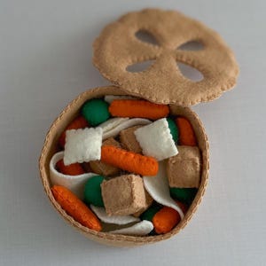 May include: A felt play food set featuring a bowl of soup with a lid. The bowl contains felt carrots, green balls, white squares, and brown cubes. The lid is a tan circle with a decorative cutout. The bowl and lid are tan.