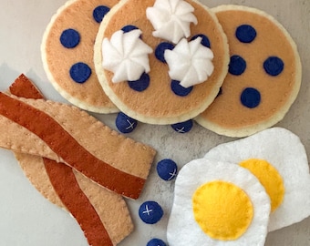 Puede incluir: Juego de desayuno de fieltro con panqueques con arándanos y crema batida, tiras de tocino y huevos fritos. Los panqueques son de color marrón claro con arándanos azul oscuro y crema batida blanca. El tocino es de color canela con rayas rojas.