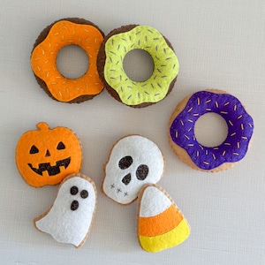 Op de afbeelding: Een set vilt Halloween-decoraties, waaronder een paars-witte donut, een groen-gele donut, een oranje pompoen, een witte geest, een witte schedel en een oranje-gele maïs.