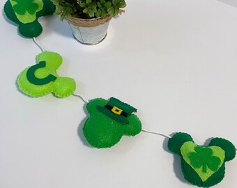 May include: A green felt garland featuring shamrock and leprechaun hat designs. The garland is strung with white thread and has a green felt shamrock with a gold buckle.