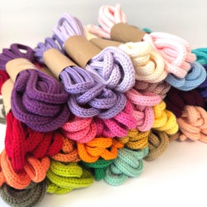 May include: A collection of colorful, braided cords in various hues, including purple, pink, green, and orange. Each cord is neatly bundled and wrapped with a brown paper band. The cords are arranged in a pile against a white background.