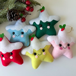May include: Five felt Christmas ornaments shaped like stars with smiling faces. Each star has a white frosting-like topping and is a different color: red, blue, green, yellow, and pink.