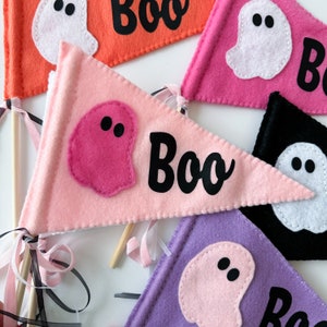 May include: Colorful felt pennant flags with a Halloween theme. Each flag features a felt ghost and the word "Boo" in black. The flags are in shades of pink, orange, purple, and black, attached to wooden dowels with ribbons.