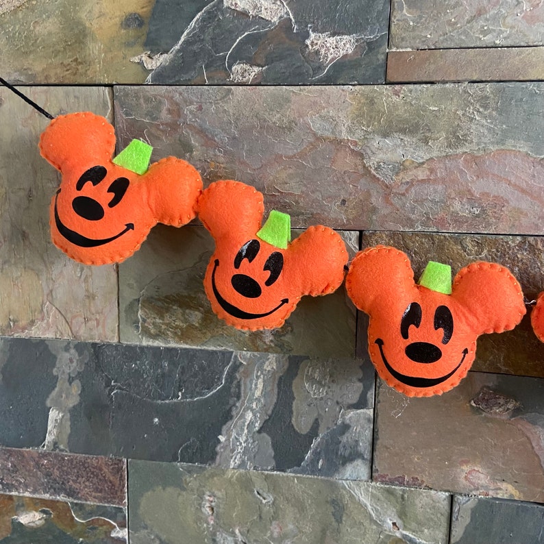 May include: A Halloween garland featuring orange felt pumpkins shaped like Mickey Mouse heads. Each pumpkin has a green felt stem and black felt eyes and nose.