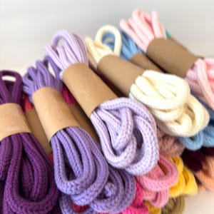 May include: A collection of thick, braided cotton cords in various colors, including purple, lavender, cream, pink, blue, yellow, and orange. Each cord is neatly folded and secured with a simple brown paper band around the center.