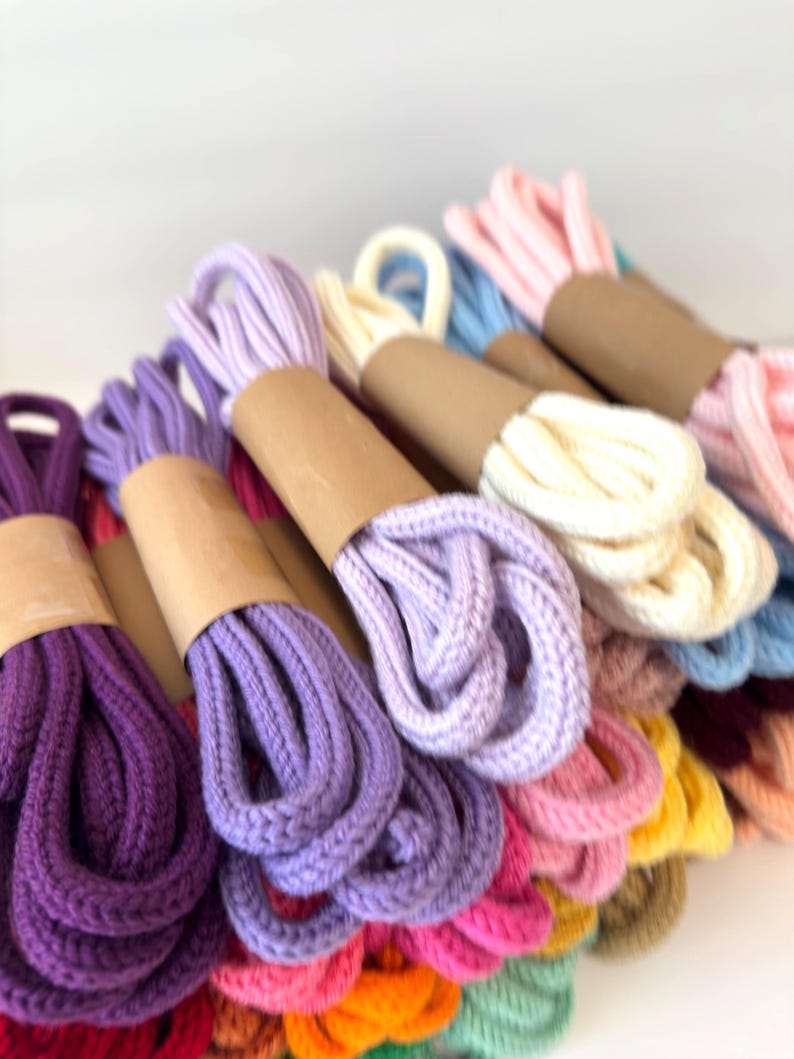 May include: A collection of thick, braided cotton cords in various colors, including purple, lavender, cream, pink, blue, yellow, and orange. Each cord is neatly folded and secured with a simple brown paper band around the center.