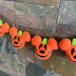 May include: A Halloween garland featuring orange felt pumpkins with Mickey Mouse ears and green stems. The pumpkins have black eyes and a black smile.