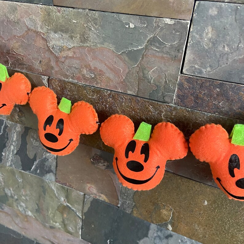 May include: A Halloween garland featuring orange felt pumpkins with Mickey Mouse ears and green stems. The pumpkins have black eyes and a black smile.