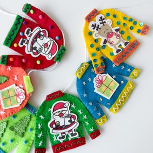 May include: A garland of felt Christmas sweaters with different designs, including Santa Claus, a reindeer, a gift, and a Christmas tree. The sweaters are strung together with white twine.