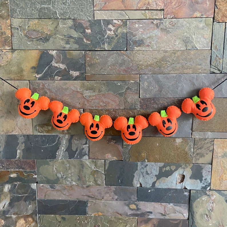 May include: A Halloween garland featuring six orange felt pumpkins with black Mickey Mouse faces and green stems. The garland is strung together with black thread and is hanging against a brick wall.