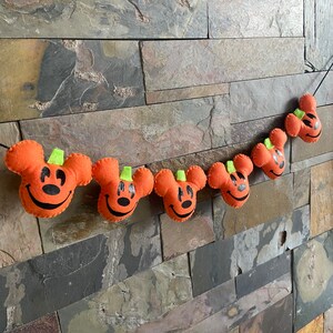 May include: A garland of orange felt pumpkins with black Mickey Mouse faces and green stems. The pumpkins are strung together with a white thread and are hanging on a brick wall.
