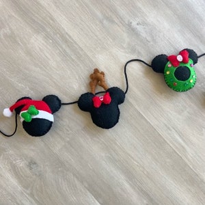 May include: A Christmas garland featuring four felt Mickey Mouse heads. The heads are decorated with a Santa hat, reindeer antlers, a red bow, and a green wreath.