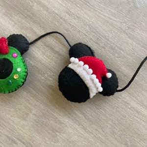 May include: A Christmas garland featuring two felt Mickey Mouse heads. One head is wearing a green wreath with red and pink accents, and the other head is wearing a red Santa hat with white trim.