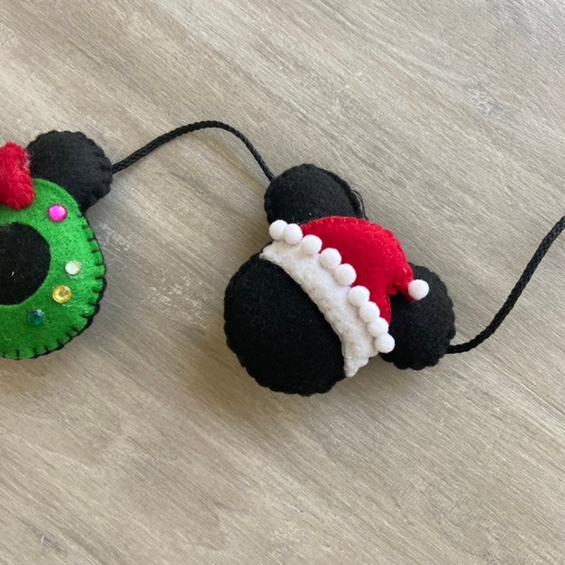 May include: A Christmas garland featuring two felt Mickey Mouse heads. One head is wearing a green wreath with red and pink accents, and the other head is wearing a red Santa hat with white trim.