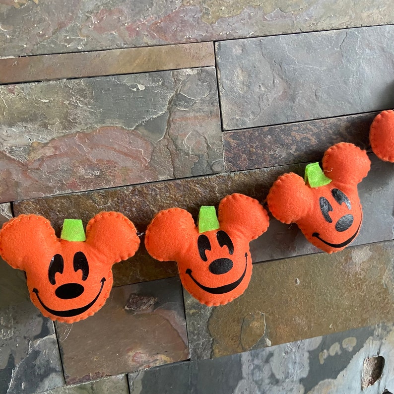 May include: A Halloween garland featuring orange felt pumpkins with Mickey Mouse faces. Each pumpkin has a green felt stem and a black felt smile.
