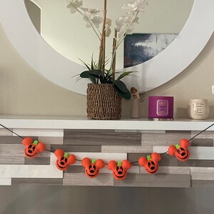 May include: A Halloween garland with orange felt pumpkins featuring a Mickey Mouse face. The garland is strung on a black cord and hangs on a wooden mantel.
