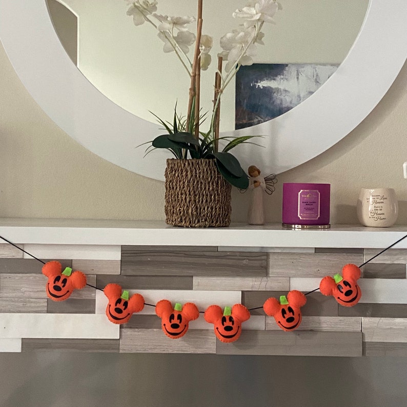 May include: A Halloween garland with orange felt pumpkins featuring a Mickey Mouse face. The garland is strung on a black cord and hangs on a wooden mantel.