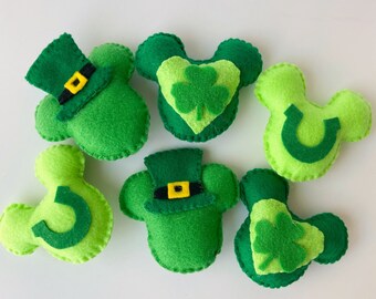 May include: Six felt green Mickey Mouse-shaped ornaments with different St. Patrick's Day themed designs. The designs include a shamrock, a horseshoe, and a leprechaun hat.