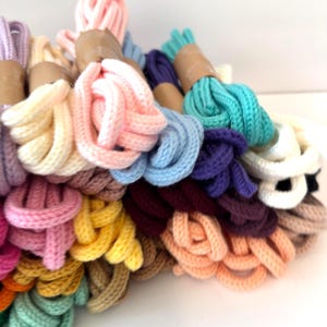 May include: A collection of colorful knitted cords in various shades, including pink, blue, yellow, and green. The cords are bundled and wrapped with brown paper. The image showcases a variety of colors and textures, suitable for crafting or decorative purposes.