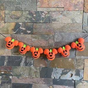 May include: A Halloween garland featuring six orange felt pumpkins, each with a black Mickey Mouse face and green stem.