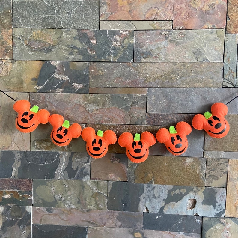 May include: A Halloween garland featuring six orange felt pumpkins, each with a black Mickey Mouse face and green stem.
