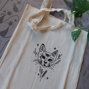 Puede incluir: Una bolsa de tela blanca con una ilustración en blanco y negro de un gato con tres ojos y un diseño floral. El gato está rodeado de estrellas.