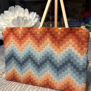 Puede incluir: Bolso de mano rectangular con un colorido patrón de chevrón. El diseño presenta filas alternas de naranja, azul claro y beige. El bolso tiene una cremallera dorada y una pequeña lengüeta de cuero.