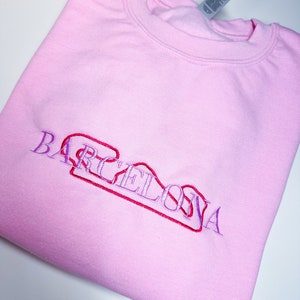 Puede incluir: Sudadera rosa con la palabra "BARCELONA" bordada en morado y rojo.