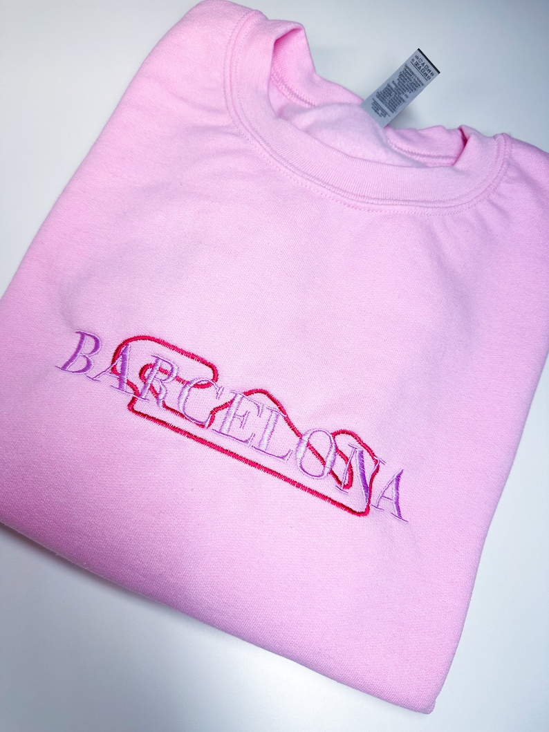 Puede incluir: Sudadera rosa con la palabra "BARCELONA" bordada en morado y rojo.