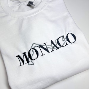 Puede incluir: Sudadera blanca con un dise&ntilde;o bordado en negro de la palabra "MONACO" con un contorno estilizado de un circuito de carreras.
