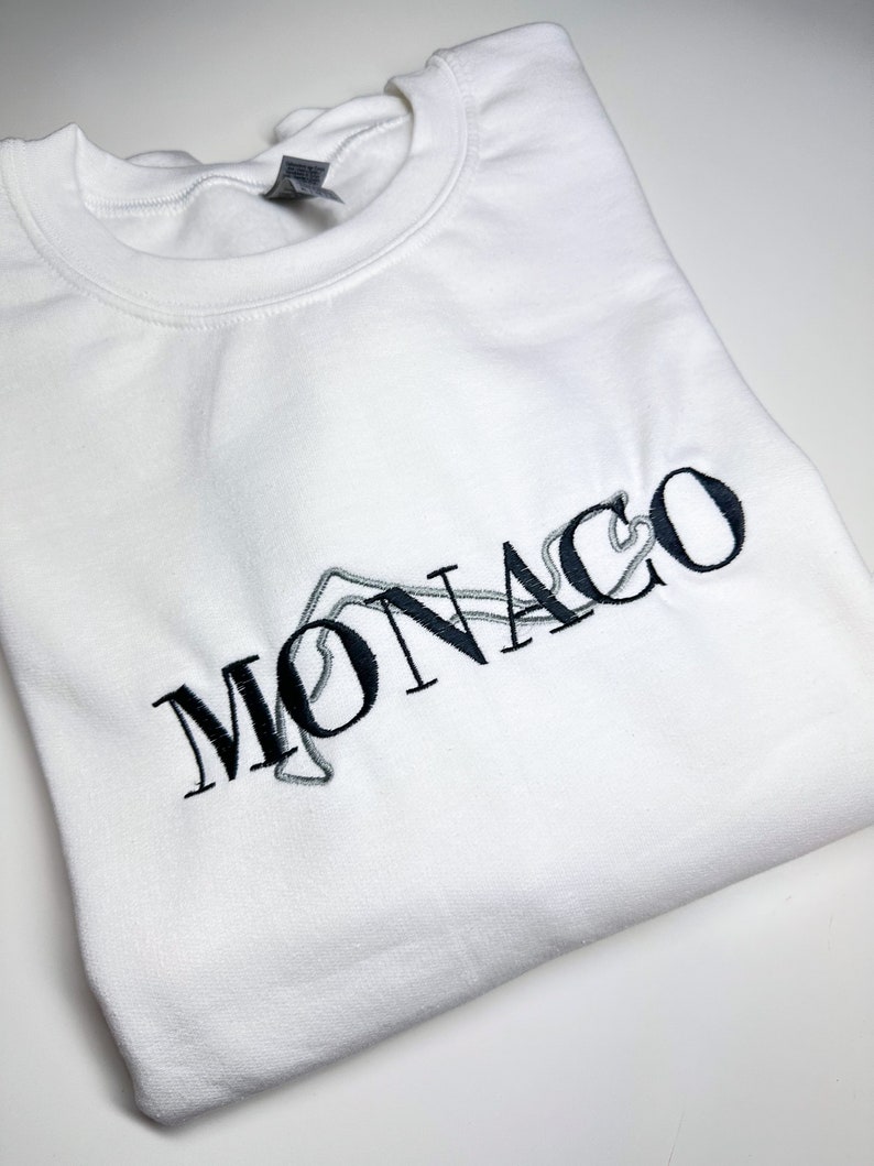 Puede incluir: Sudadera blanca con un dise&ntilde;o bordado en negro de la palabra "MONACO" con un contorno estilizado de un circuito de carreras.