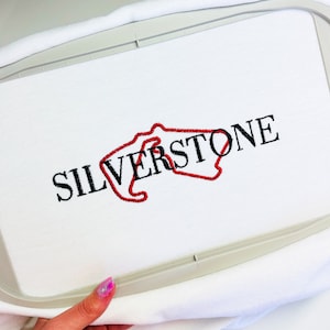Puede incluir: Tela blanca con un contorno bordado en rojo del circuito de Silverstone y la palabra "SILVERSTONE" en bordado negro.