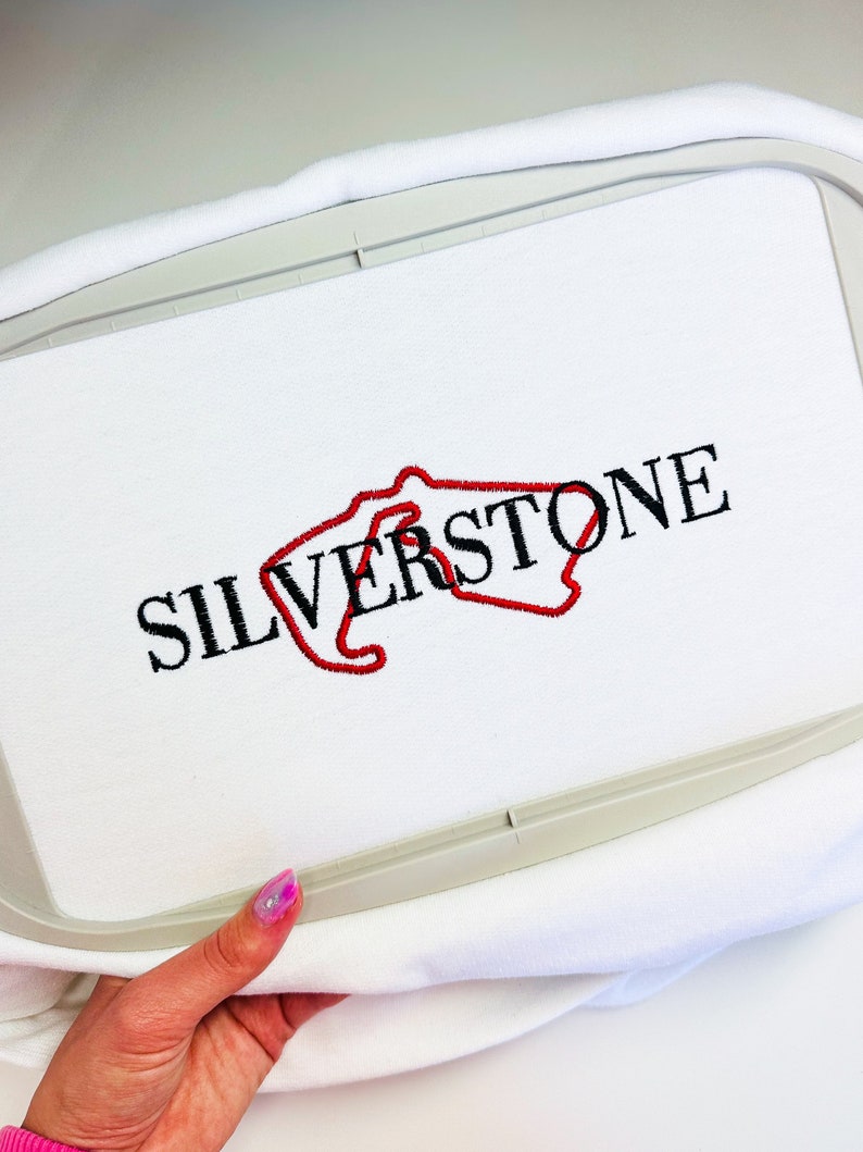Puede incluir: Tela blanca con un contorno bordado en rojo del circuito de Silverstone y la palabra "SILVERSTONE" en bordado negro.