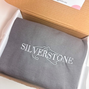 Puede incluir: Una sudadera gris con un dise&ntilde;o bordado en blanco del circuito de Silverstone y la palabra "SILVERSTONE".