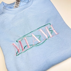 Puede incluir: Una sudadera azul claro con la palabra "MIAMI" bordada en rosa, verde y blanco.