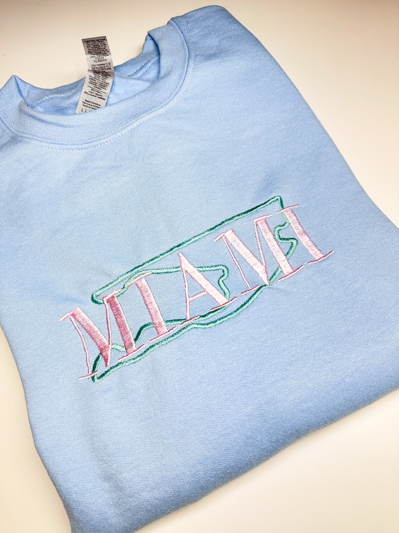 Puede incluir: Una sudadera azul claro con la palabra "MIAMI" bordada en rosa, verde y blanco.