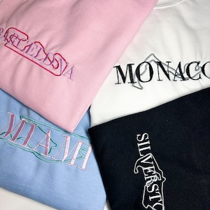Könnte beinhalten: Vier Sweatshirts mit gestickten Städtenamen und Strecken-Designs. Die Sweatshirts sind rosa, weiß, hellblau und schwarz. Die Namen sind Barcelona, Monaco, Miami und Silverstone.