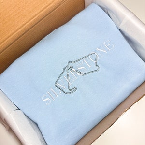 Puede incluir: Una sudadera de color azul claro con la palabra "SILVERSTONE" bordada en blanco en la parte delantera.