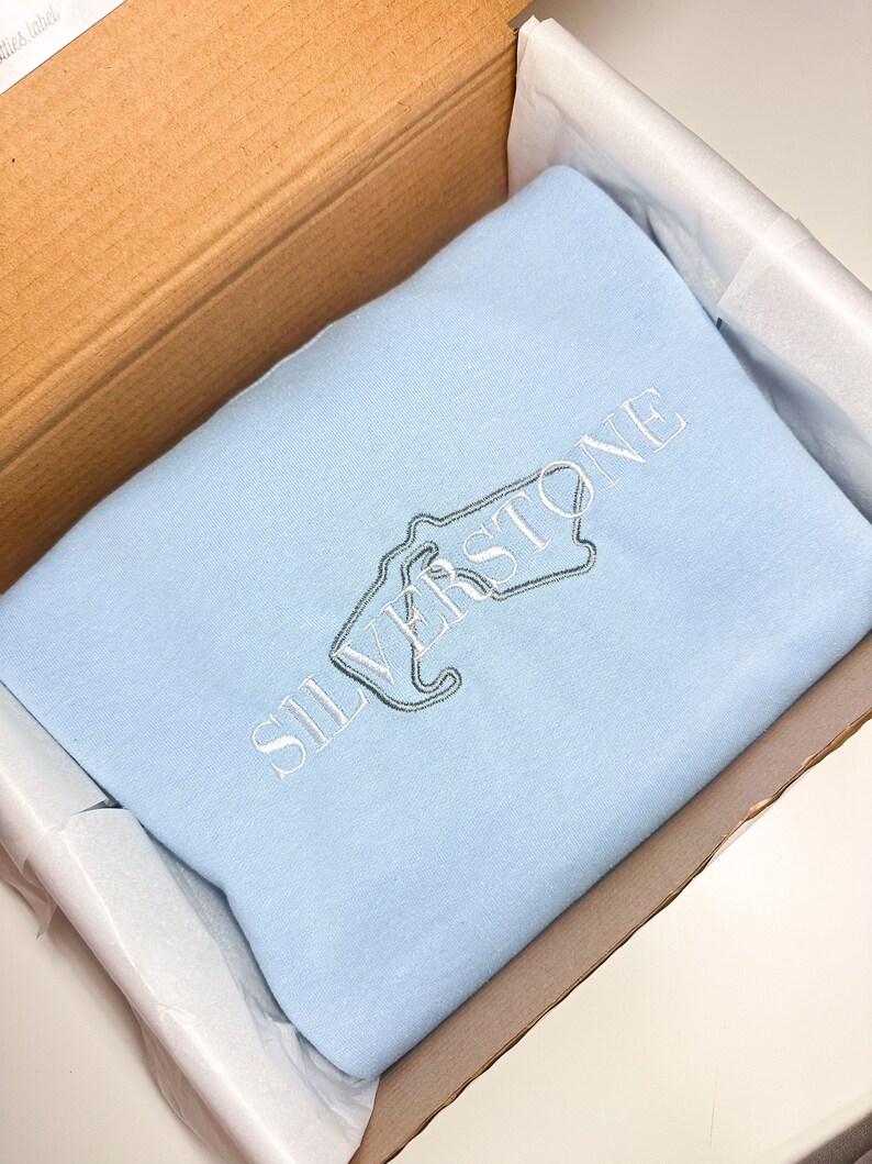 Puede incluir: Una sudadera de color azul claro con la palabra "SILVERSTONE" bordada en blanco en la parte delantera.